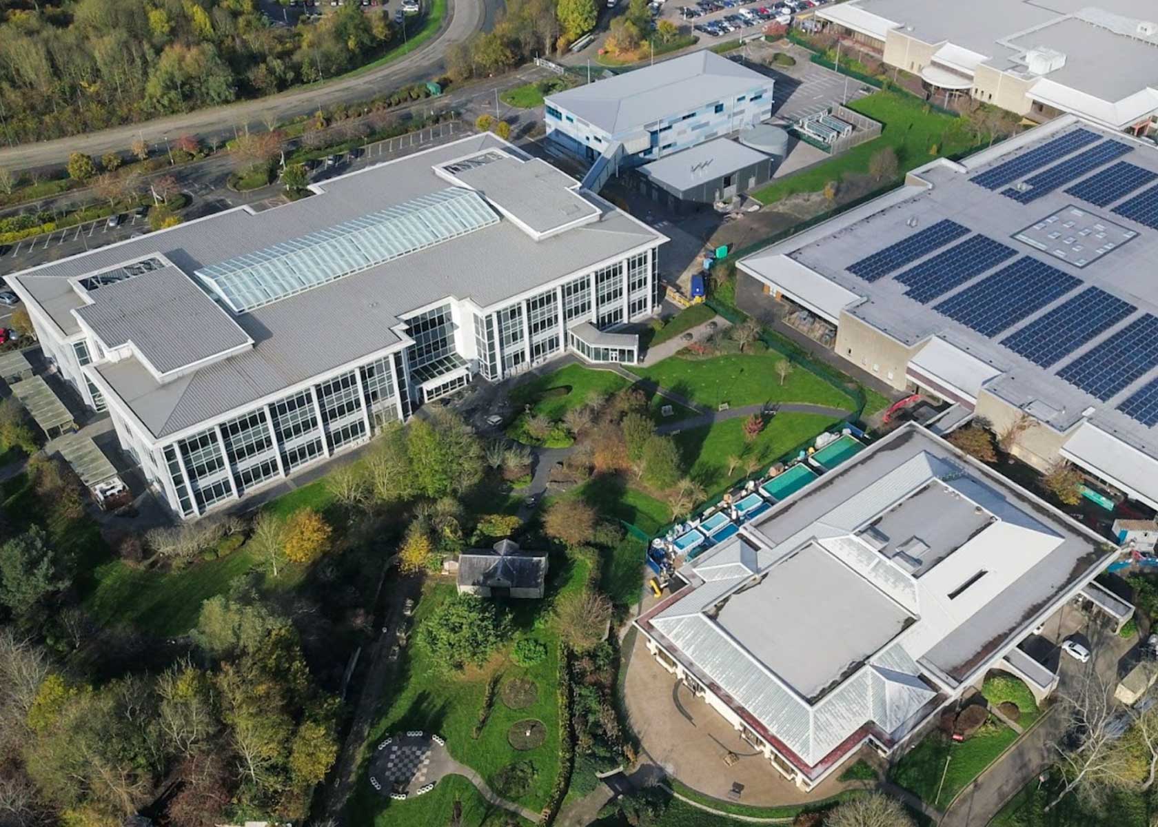 Aerial view of a commercial or institutional complex with large rooftop solar panel installations on multiple buildings, surrounded by greenery and parking areas.