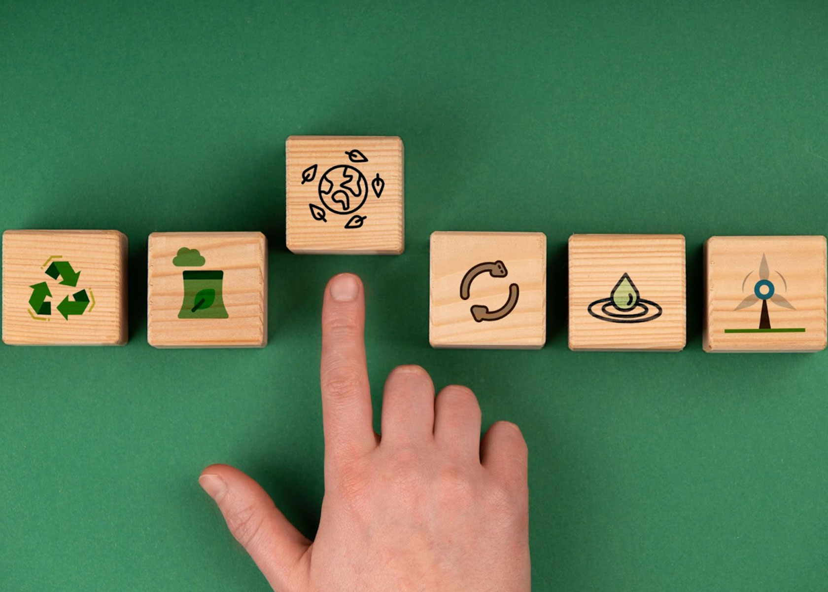 A hand pointing at a wooden block with a sustainability icon of the Earth surrounded by leaves, placed among other blocks depicting symbols of recycling, renewable energy, clean water, and eco-friendly practices on a green background.