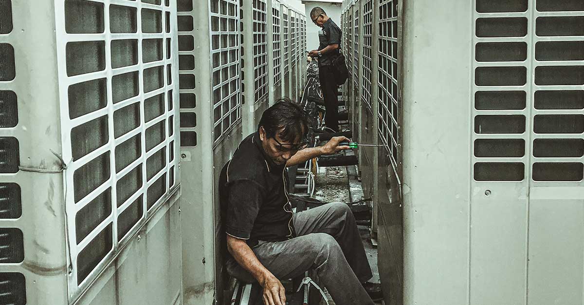 Technicians performing maintenance work between large industrial heat pump or HVAC outdoor units installed on a building rooftop.